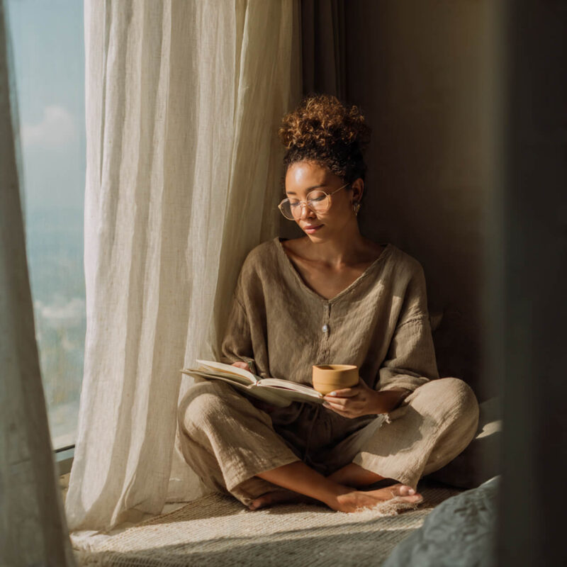 Calm person journaling by a sunlit window with a cup of tea, wearing neutral tones, symbolising peace, reflection, and journaling for overthinking