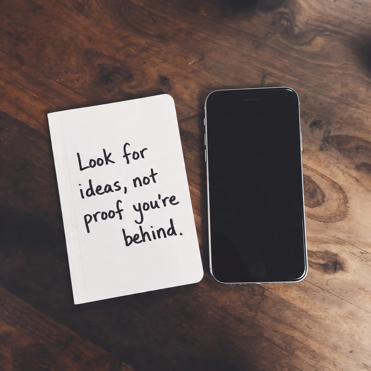 Notebook beside a smartphone on a wooden table, handwritten note reads “Look for ideas, not proof you’re behind,” illustrating realistic self-talk and bridge affirmation practice.