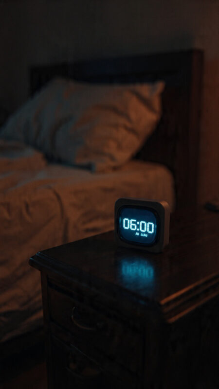 06:00 alarm glowing on a bedside table, marking consistent sleep and start–stop cut-offs.