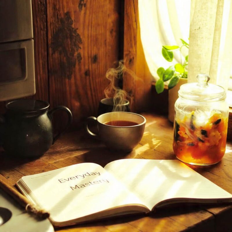 “Cup of tea beside a jar of sauerkraut and an open notebook titled ‘Everyday Mastery’ on a wooden kitchen counter in warm morning light, symbolising calm gut health rituals.”