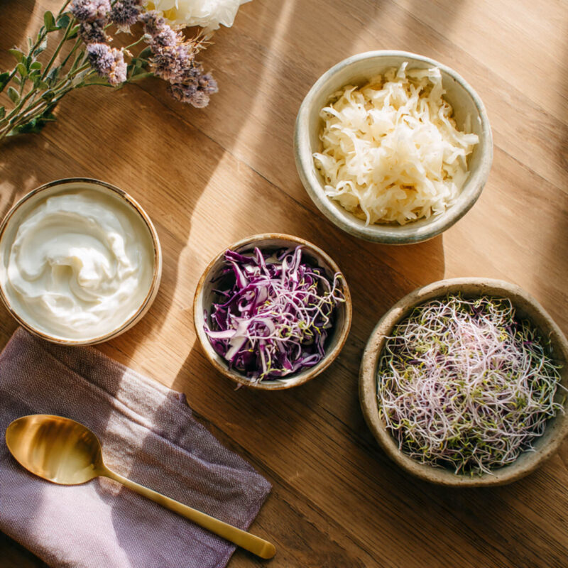 Flat lay of probiotic foods including sauerkraut, Greek yogurt, and microgreens on a wooden table with lavender cloth and gold spoon, illustrating simple gut health habits