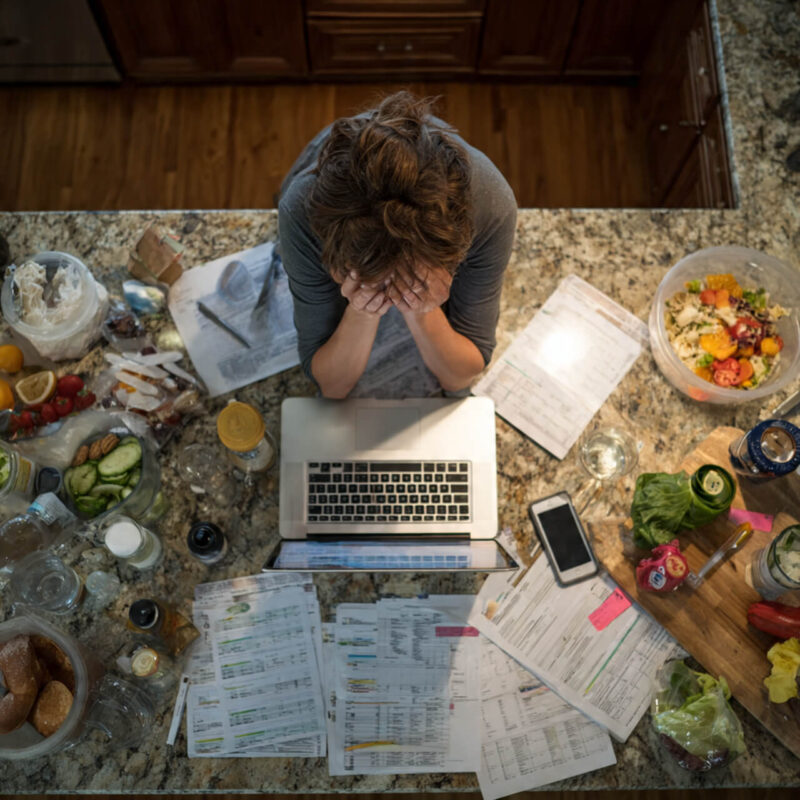Overhead view of parent surrounded by tasks at kitchen counter — laptop, grocery lists, and children’s papers — showing the invisible mental load behind decision fatigue
