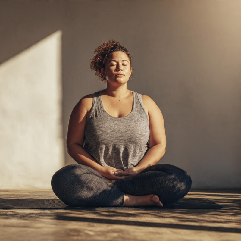Overweight person sitting cross-legged on a yoga mat, eyes closed and breathing calmly, representing reflection and self-respect at the start of a fitness journey.