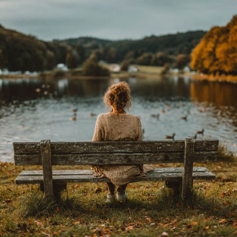 Person sitting on a wooden park bench beside a lake, watching ducks in autumn light, symbolising calm rest and reflection.