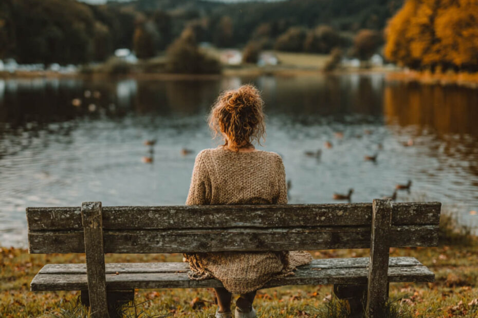 Person sitting on a wooden park bench beside a lake, watching ducks in autumn light, symbolising calm rest and reflection.