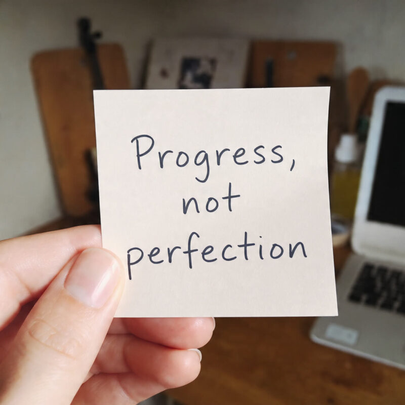 Hand holding a sticky note reading “Progress, not perfection,” with a blurred desk in the background, symbolising self-compassion and realistic growth