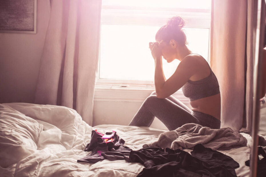 Woman sitting on edge of unmade bed in workout clothes struggling with motivation in early morning light
