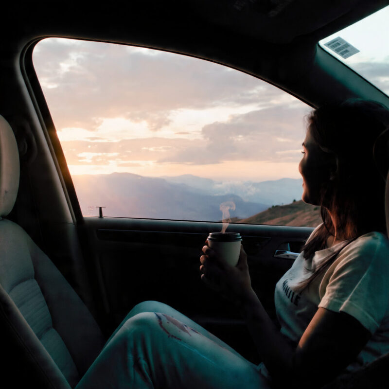 A person sits inside a car holding a cup of coffee, watching the sunrise over distant hills — capturing the peace and presence of simple micro-adventures that help you feel alive again.