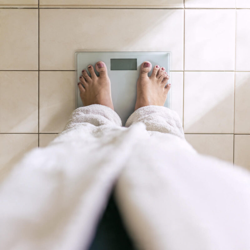 Person in white bathrobe standing on bathroom scale Monday morning weigh-in
