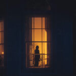 Laziness Is a Lie - Here's What's Really Keeping You Comfortable Woman standing at window at dusk contemplating change - representing the difference between laziness and burnout caused by old identity wiring