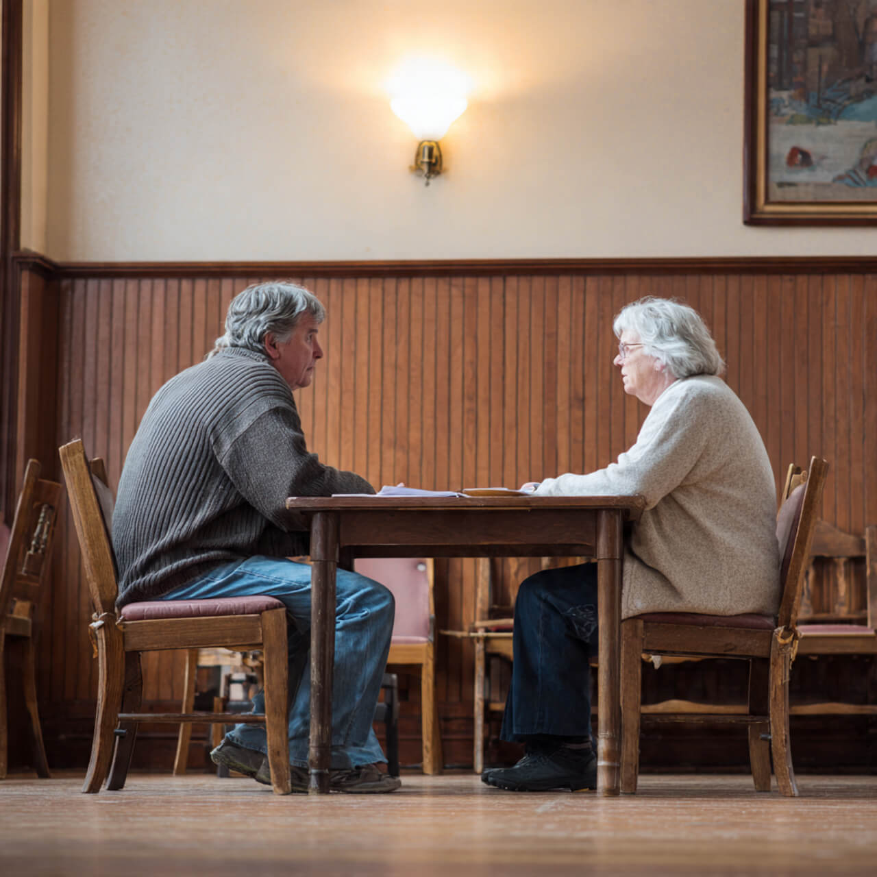Two people sitting across from each other at a wooden table in a community hall, having a warm volunteering conversation that reduces loneliness and symbolises volunteer work for loneliness