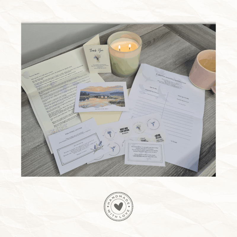 The Stoic Letter monthly subscription pack laid out on grey wood surface including handwritten letter, collectible postcard with sunset bridge artwork, gentle reflection worksheet, breathing practice card, cream envelope and pocket tools alongside a lit candle and pink mug of tea